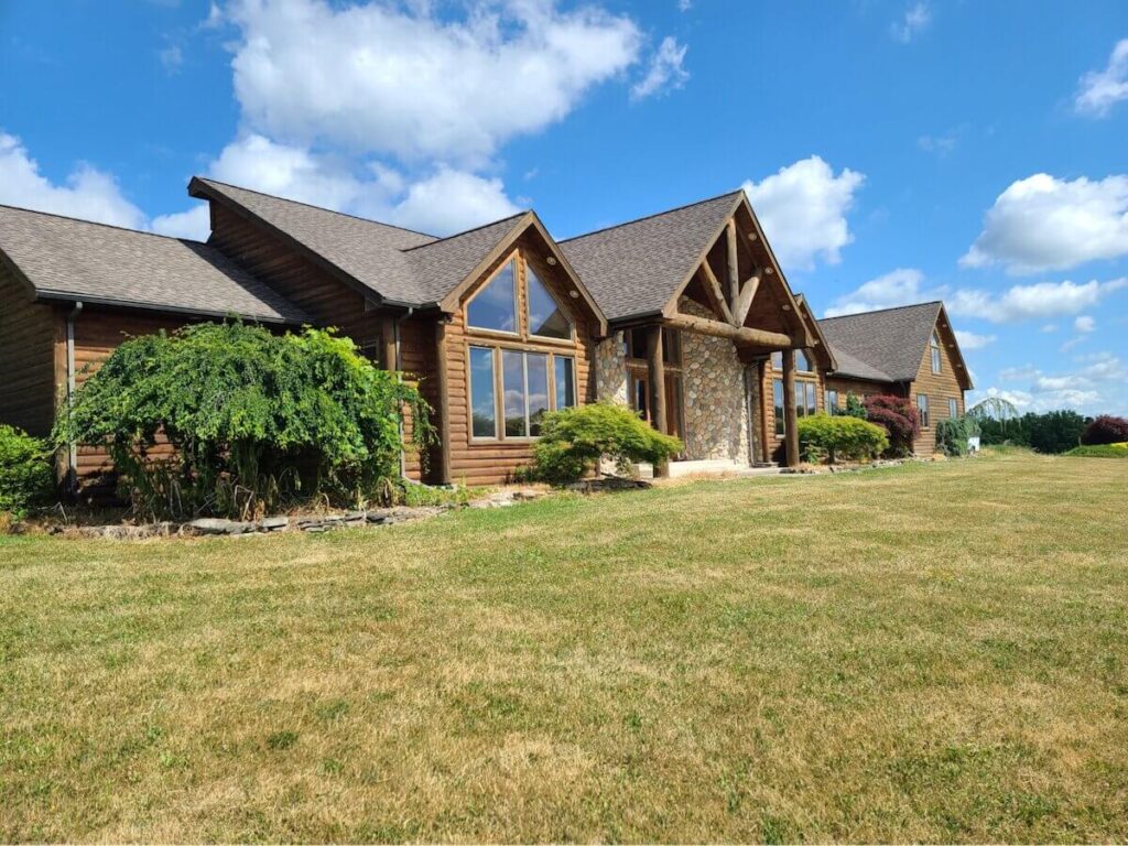 Lodge-style home with TruLog Western Cedar steel log siding, a stone chimney, exposed wood beam gable, and large front windows against a blue sky.