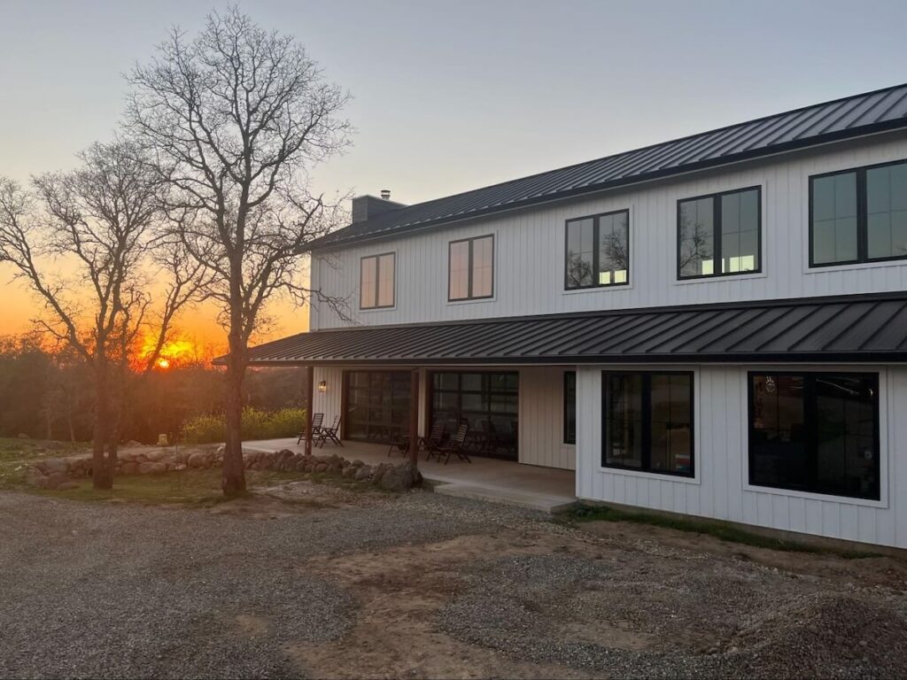 Two-story barndominium with white TruLog steel siding, a black metal roof, and black trim at sunset.