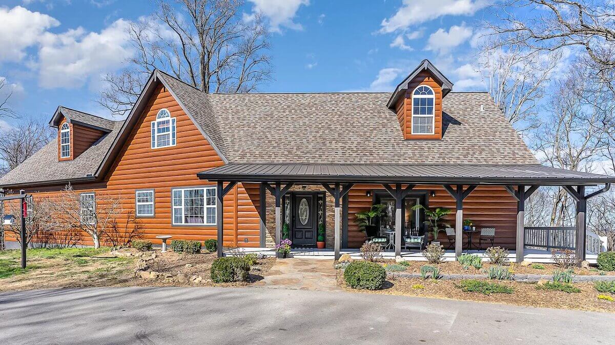 Home with TruLog steel siding, a covered front porch with stone columns, and dormer windows on a wooded lot.