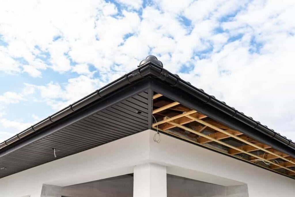 Corner of a house with black vented soffit panels, gutters, and fascia, with exposed wooden rafters visible where soffit installation is incomplete.