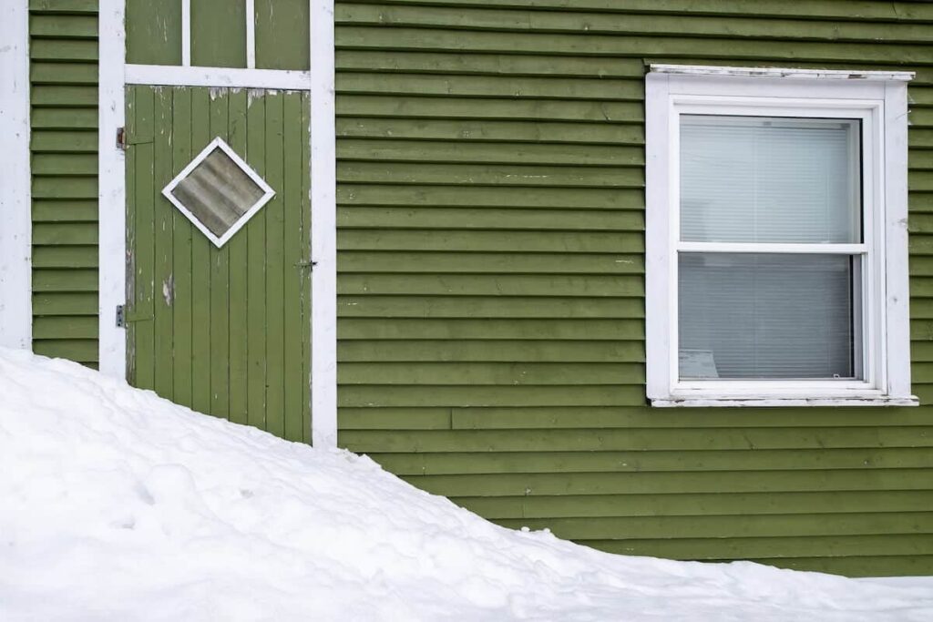 The side of a home with a snowdrift against its green wood siding