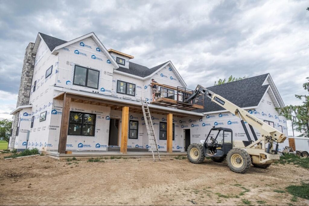 A person stands on a telehandler platform to install siding on the second story of a new home