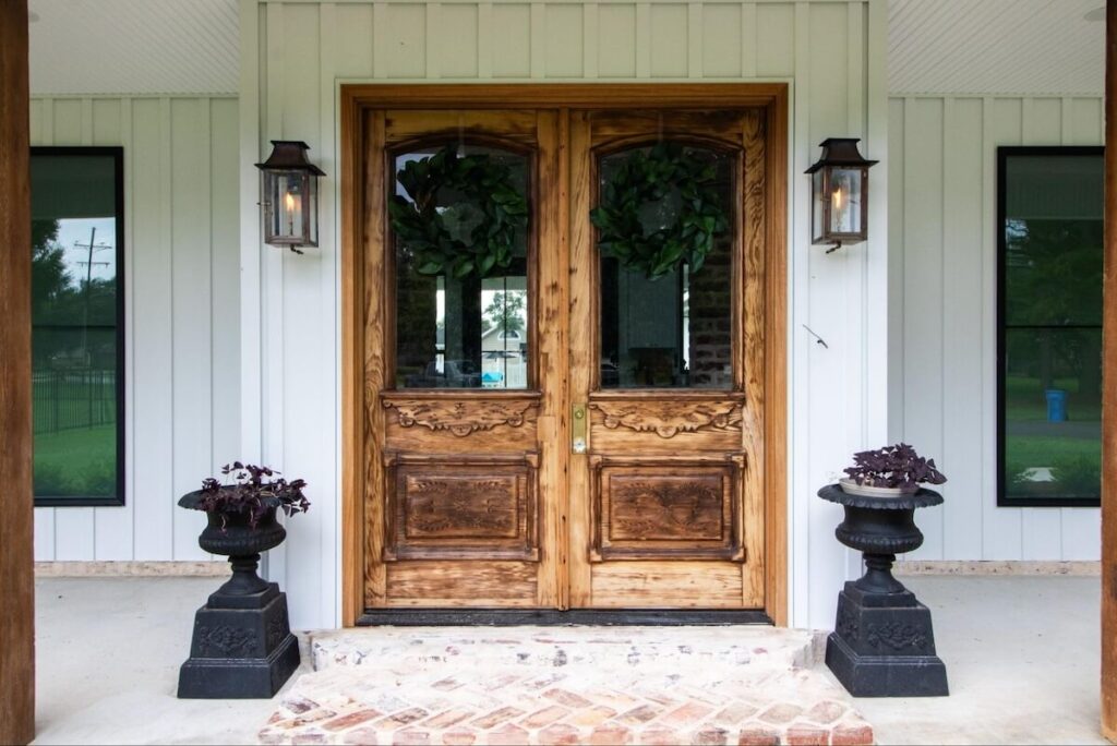 A closeup of a front porch with upgraded lighting, a wide ornate wooden door, and TruLog steel board and batten siding in Matte White