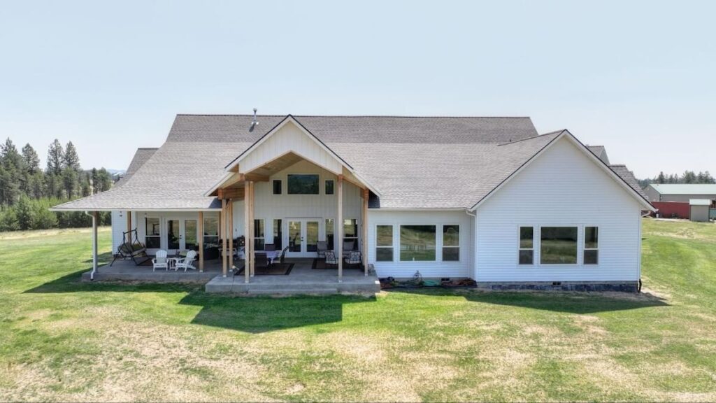 A large covered patio attached to a white farmhouse-style home overlooking an open grassy yard