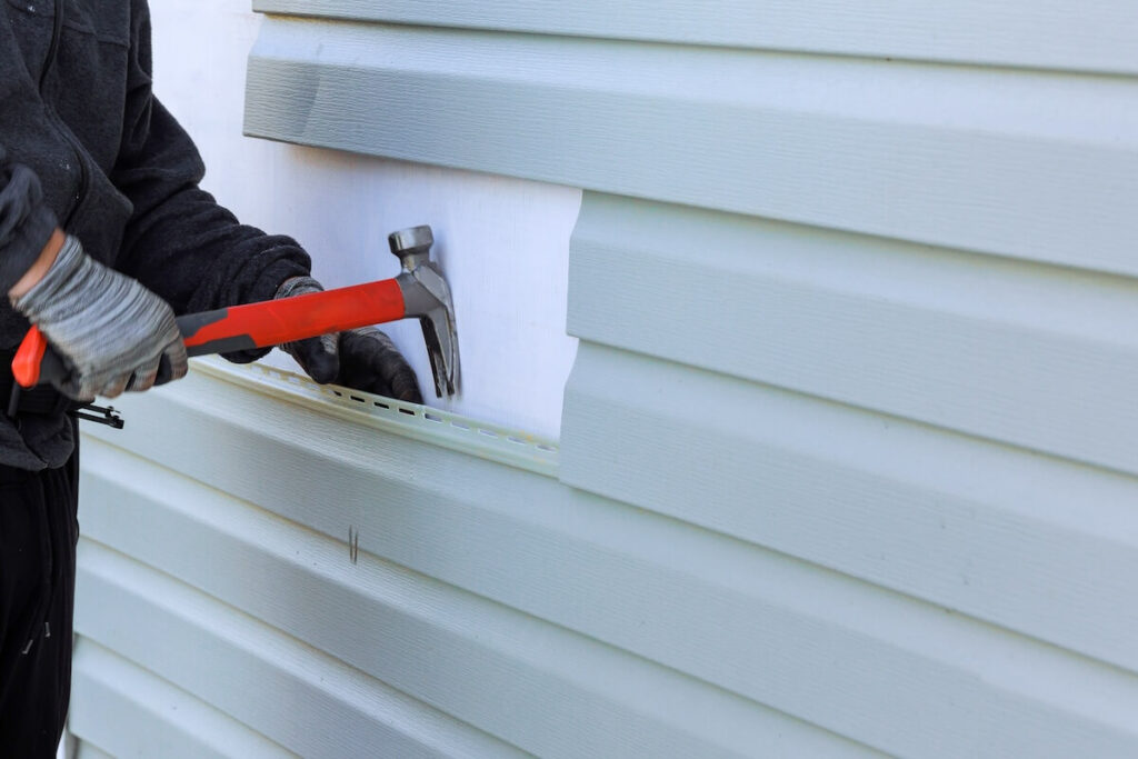 Contractor using a claw hammer to remove and replace a section of light gray vinyl siding.