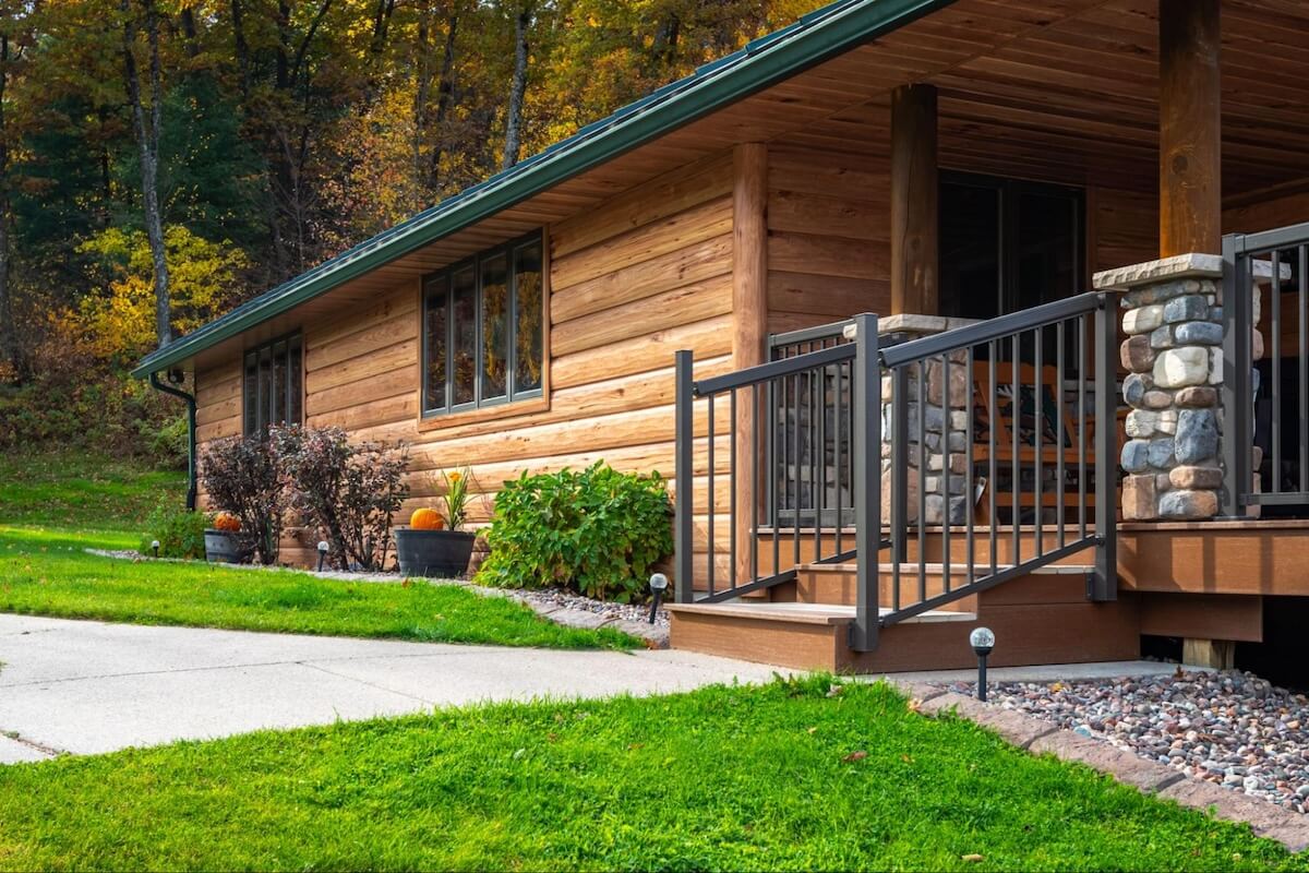 Cabin-style home with warm-toned TruLog steel siding with realistic wood grain, stone porch columns, and a green metal roof surrounded by autumn foliage.