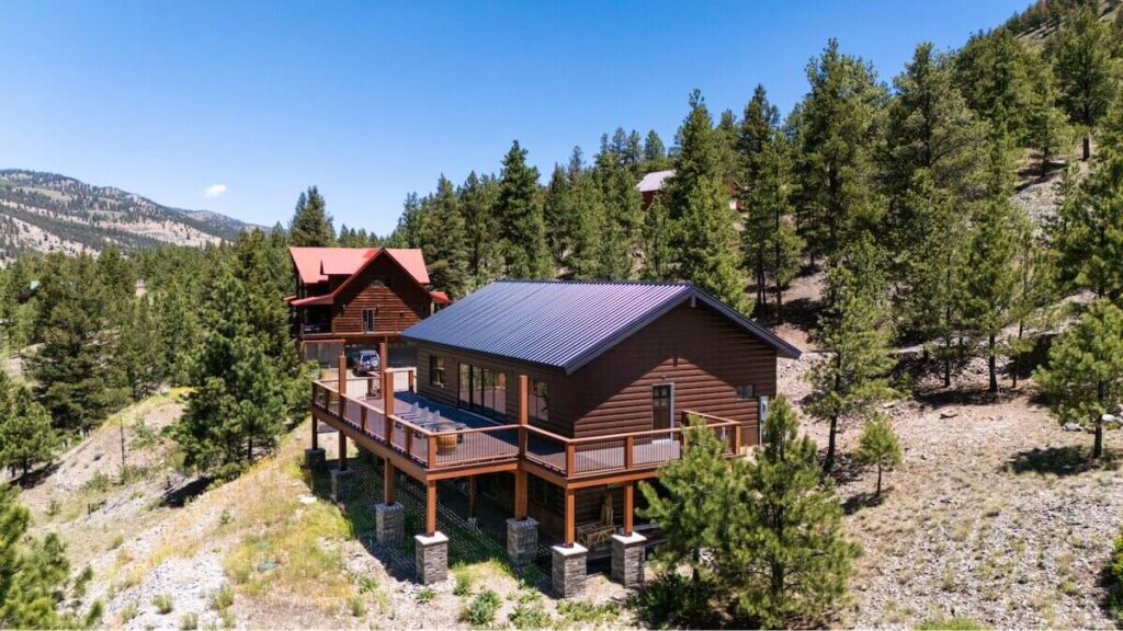Aerial view of a mountain home with dark brown steel log siding and a metal roof surrounded by pine trees on a hillside