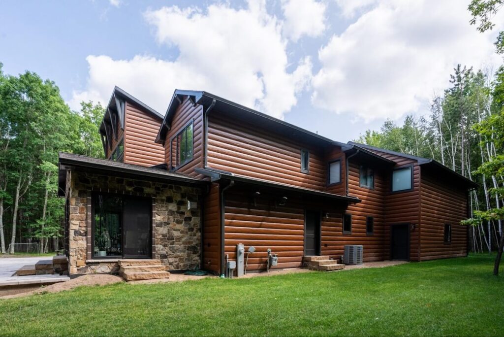 Two-story home with rich brown steel log siding and natural stone detailing set against a forested backdrop.