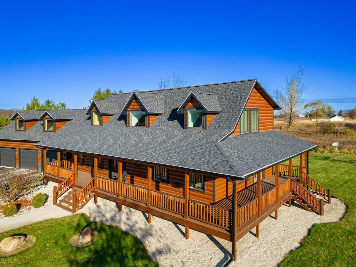 Aerial view of a large home with warm golden TruLog steel siding, a dark shingle roof, a wraparound deck, and a manicured yard, set against a clear blue sky.