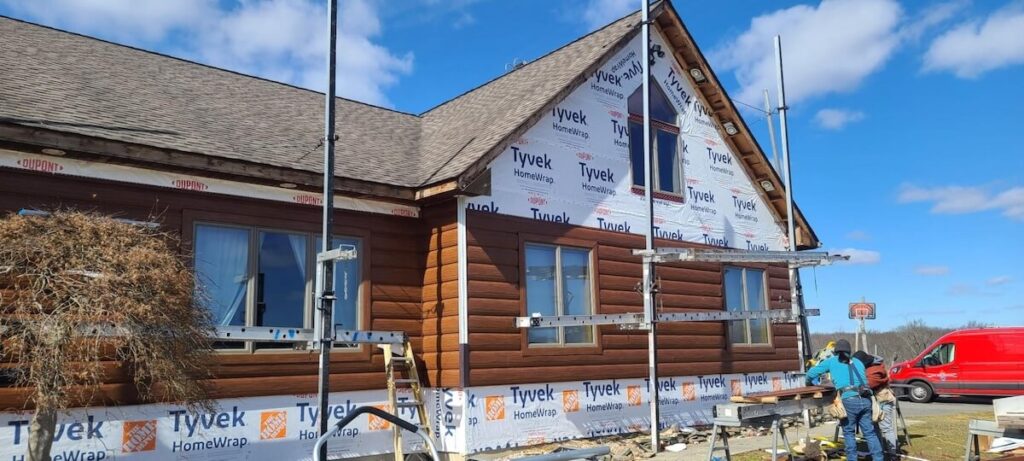 A log home mid-remodel with warm brown TruLog steel siding being installed over house wrap with workers on-site on a clear day.