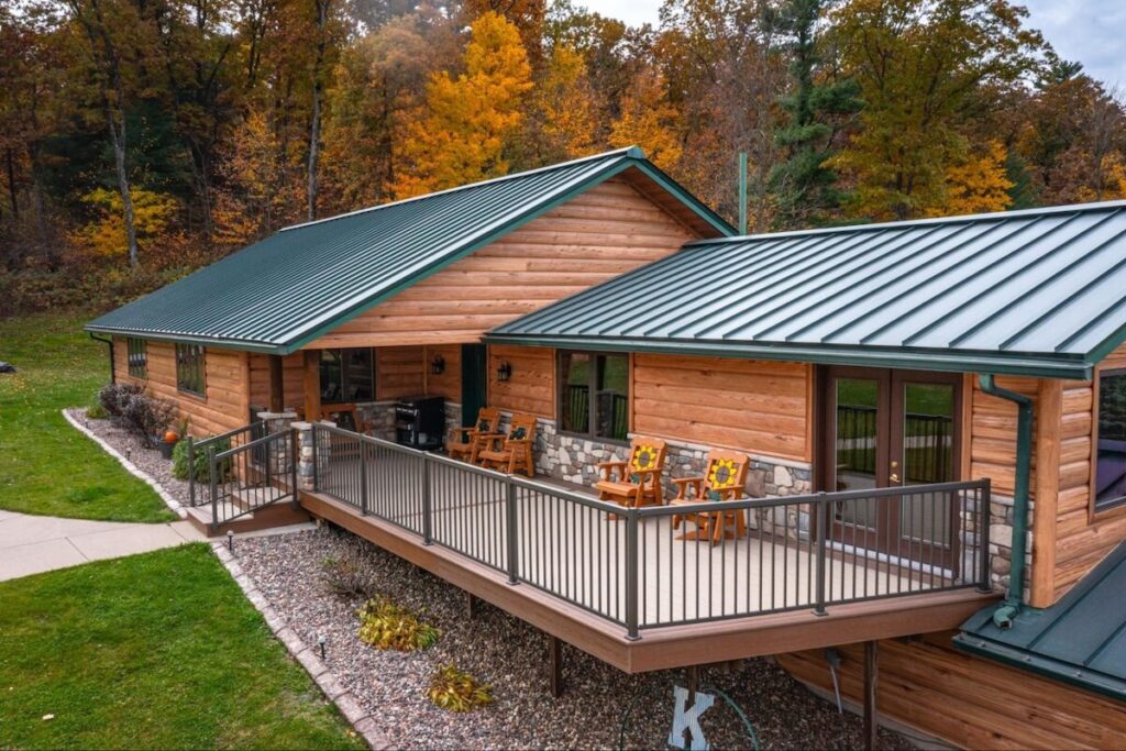 Log cabin with TruLog steel siding and a green standing-seam metal roof, featuring a spacious deck and stone accents, surrounded by autumn foliage.