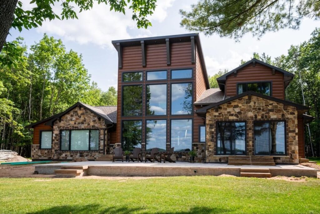 Modern log home with rich brown TruLog siding, natural stone accents, dark trim details, and large windows, nestled against a forest.