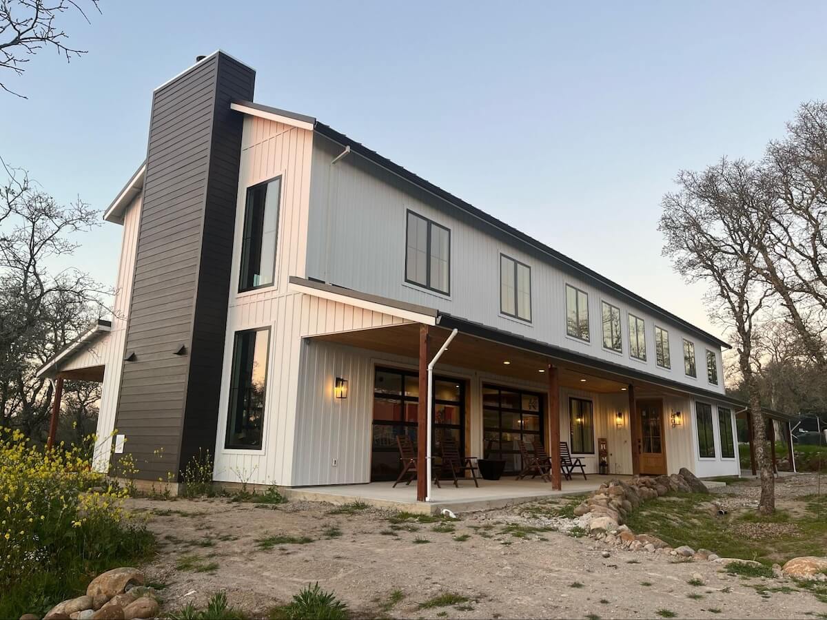 Two-story barndominium with white vertical TruLog steel siding, dark accent panels, large glass garage doors opening to a covered patio with wood timber porch supports.