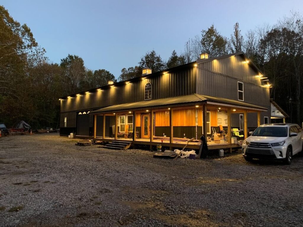 Dark gray two-story barndominium with vertical TruLog metal siding, warm exterior lighting along the roofline, and a wraparound wood porch at dusk.
