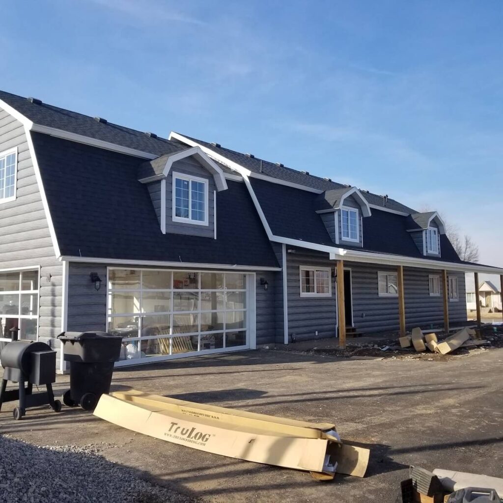 Cape Cod-style home with gray TruLog steel lap siding, white trim, dormer windows, and a glass-panel garage door set against a blue sky.
