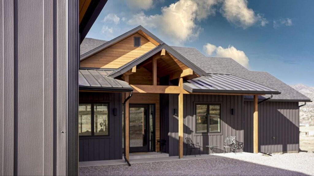 Close-up view of a farmhouse-style home featuring dark gray TruLog siding, natural wood gable accents and porch beams, a standing-seam metal roof, and dark-framed windows in a mountain setting.