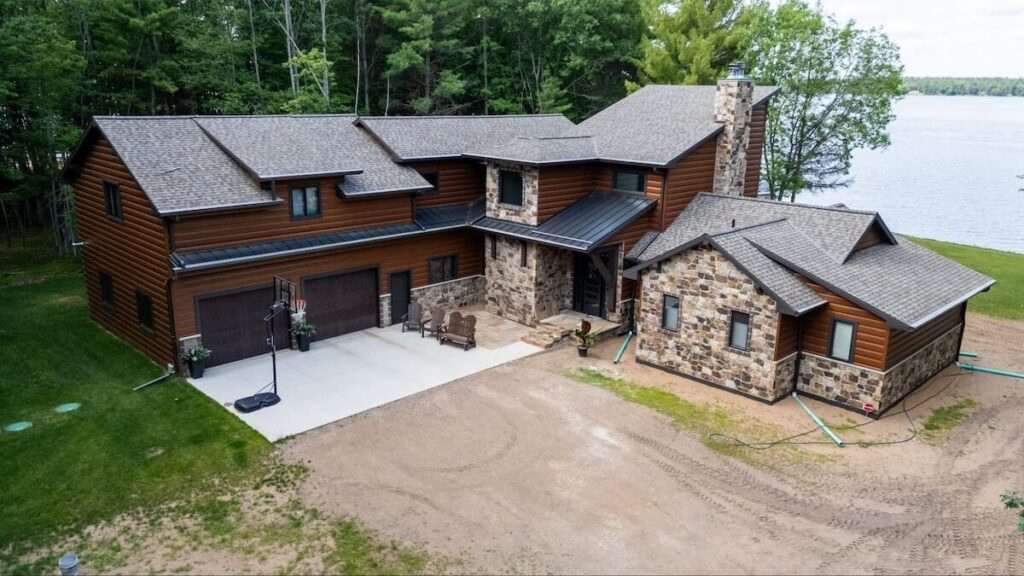 Aerial view of a lakeside home with dark brown TruLog siding, natural stone accents, and a multi-gable gray shingle roof.