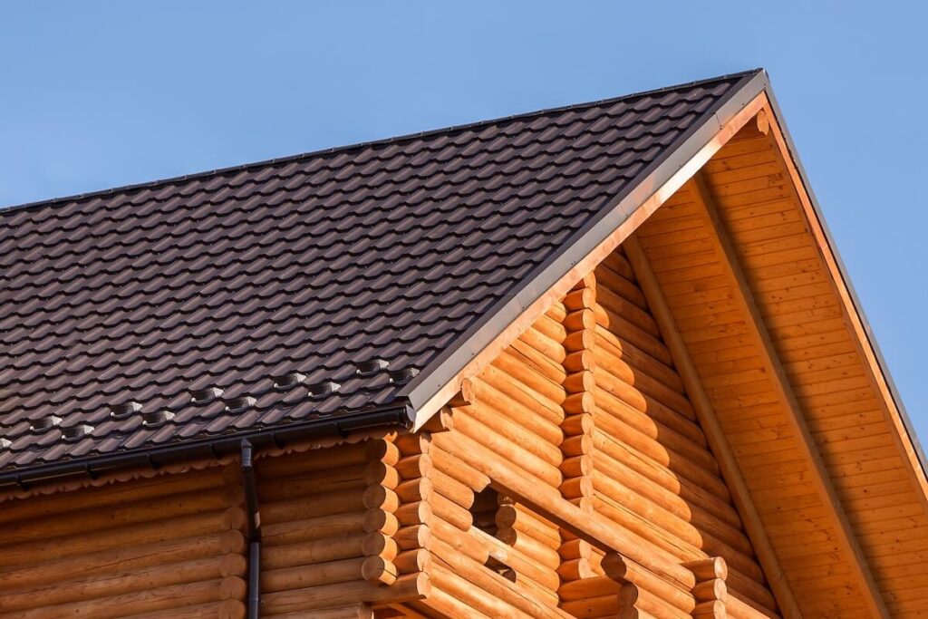 Gable of a log cabin with stacked round log siding and a dark tile roof against a clear blue sky.