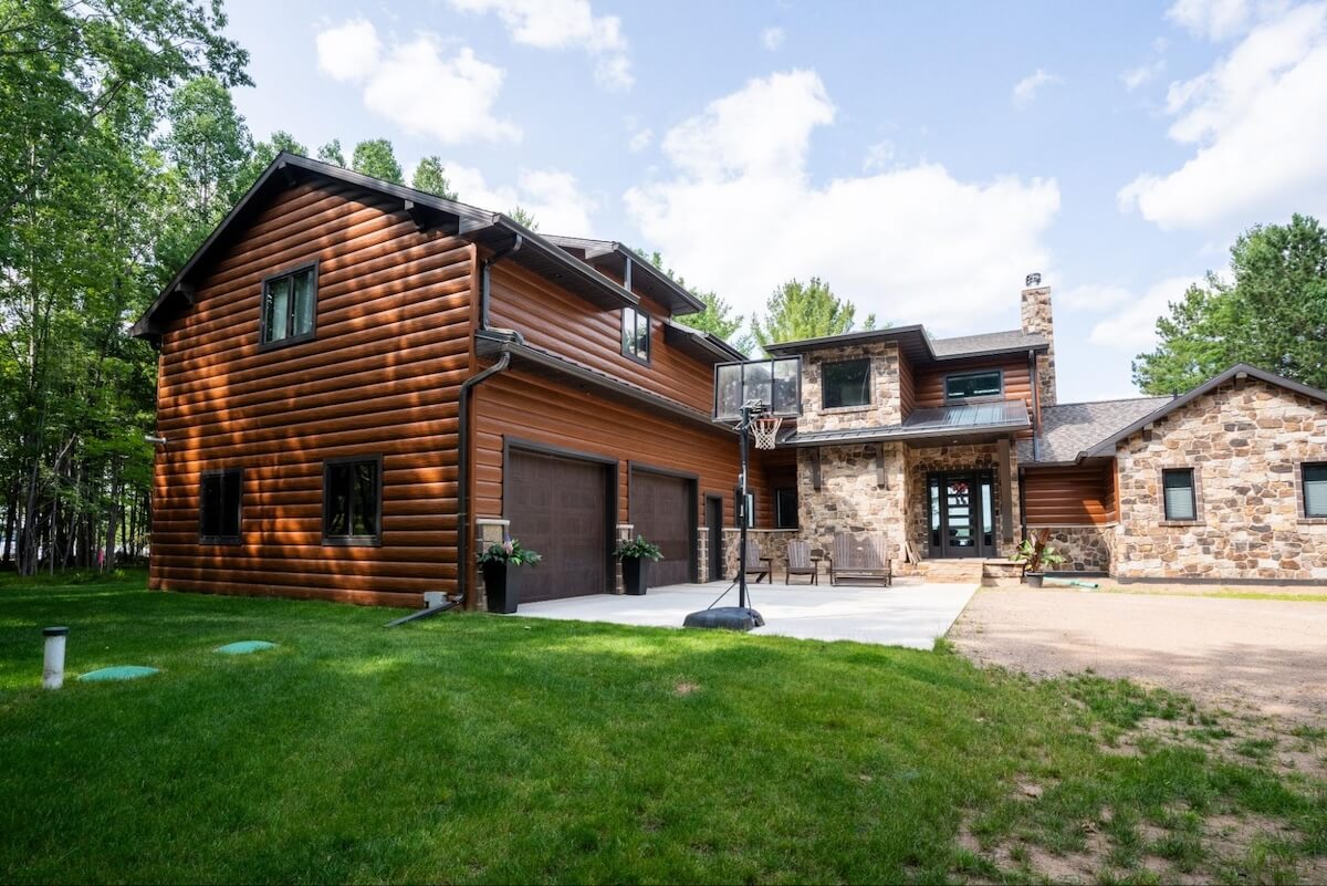 Multi-level home featuring rich brown TruLog steel siding panels with natural stone accents, matching brown garage doors, and a landscaped concrete driveway.