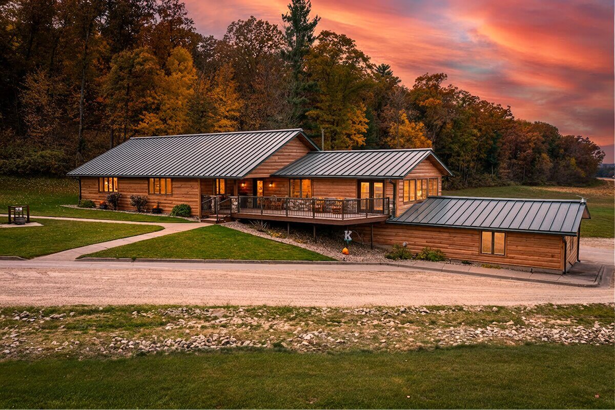Ranch-style home with TruLog steel log look siding and a standing-seam metal roof, set against a wooded backdrop with fall foliage at sunset.