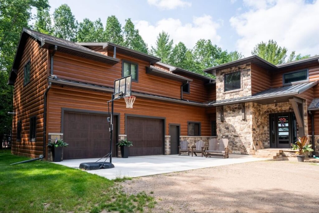 Two-story home with brown TruLog steel log look siding and matching wood-tone garage doors, paired with natural stone accents around the entryway, surrounded by mature green trees.