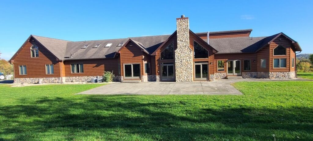 Sprawling two-story home with warm-toned TruLog synthetic log siding, a river stone chimney, and a dark shingled roofline set on a manicured green lawn under a clear blue sky.