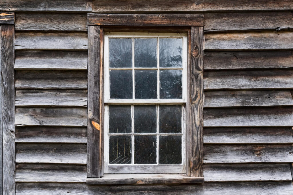 Close-up of weathered gray barnwood siding on an old structure, showing aged wood grain, natural patina, and a rustic window frame.