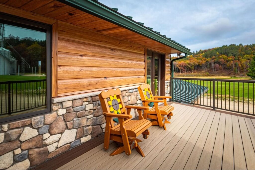 Covered deck of a cabin with TruLog steel log siding, stone veneer accents, green metal roof with visible fascia, and wooden Adirondack chairs overlooking a rural autumn landscape.