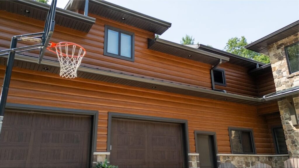 Two-story home with warm wood-tone TruLog steel log siding, dark fascia and gutters, stone accents, brown garage doors, and a wall-mounted basketball hoop.