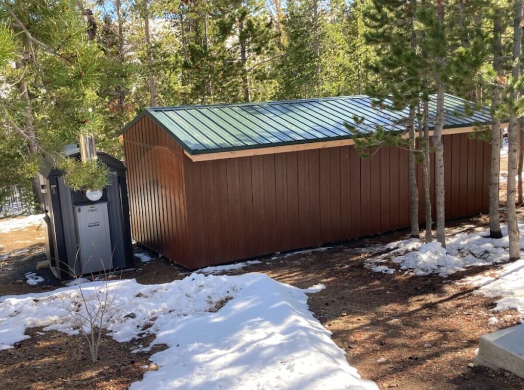 TruLog steel siding in a dark brown woodgrain finish on a utility shed with a green metal roof, surrounded by pine trees.