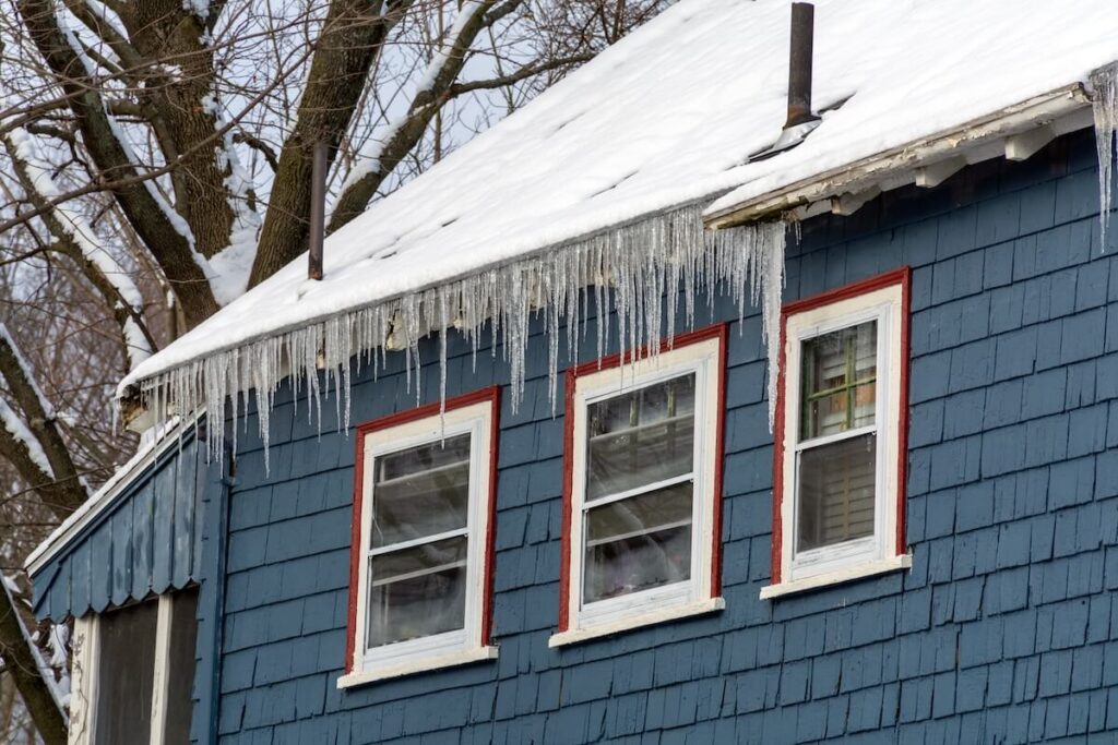 Exterior of a house with blue painted cedar shake siding and a snow-covered roof with icicles hanging from the eaves.