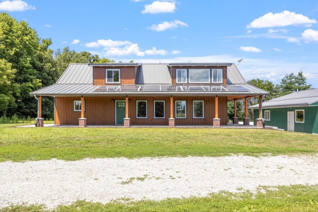 Brown board-and-batten TruLog steel siding home with a standing seam metal roof, wraparound porch, and rooftop solar panels