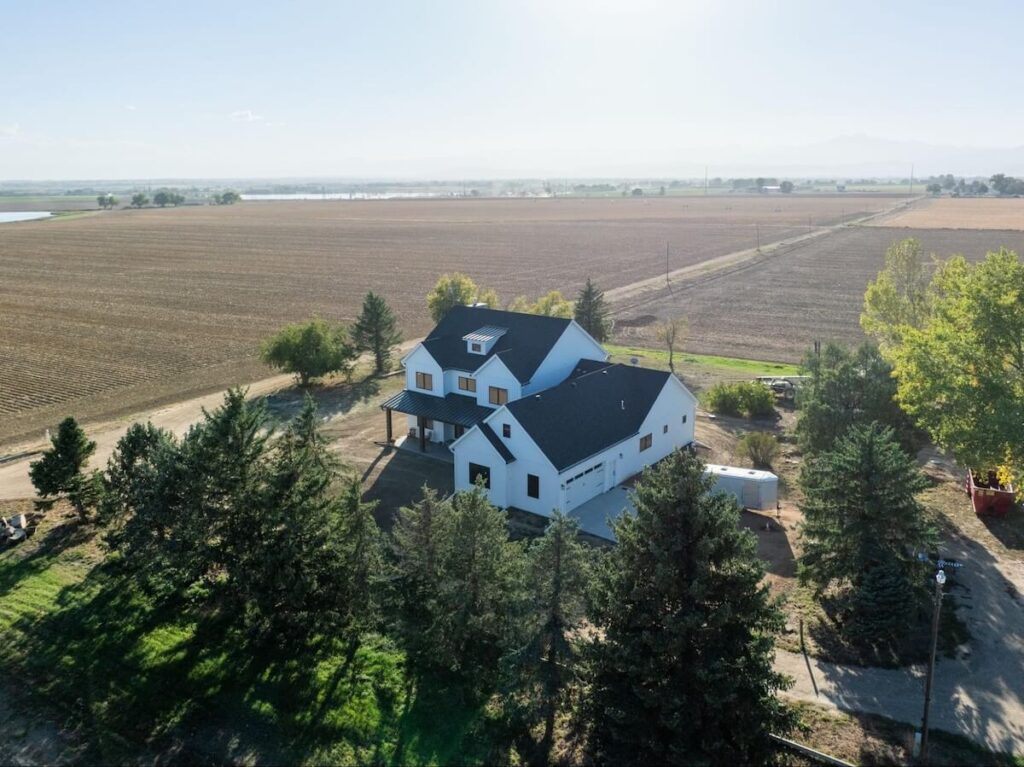 Aerial view of a white farmhouse with black metal roofing and vertical TruLog steel siding, surrounded by trees and open agricultural fields.